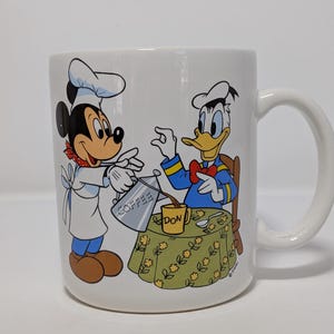 May include: White ceramic mug featuring Mickey Mouse as a chef pouring coffee for Donald Duck. The mug has a handle and a colorful illustration of the Disney characters. The table has a floral pattern and the word "DON" is visible.
