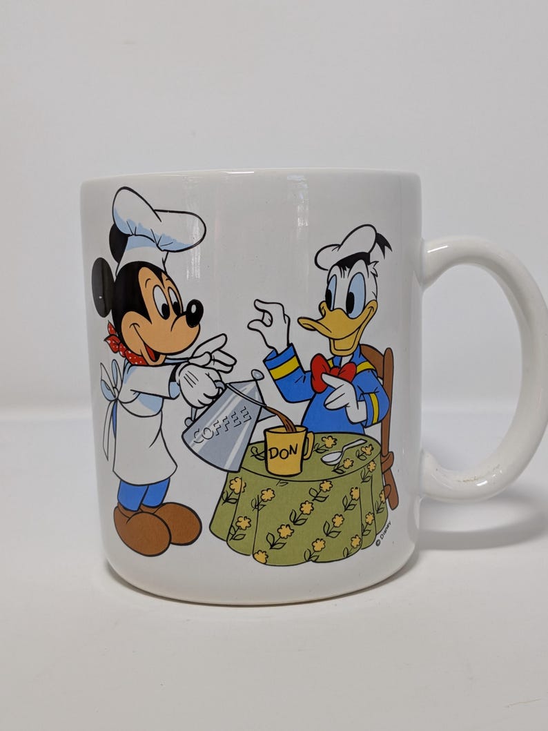 May include: White ceramic mug featuring Mickey Mouse as a chef pouring coffee for Donald Duck. The mug has a handle and a colorful illustration of the Disney characters. The table has a floral pattern and the word "DON" is visible.
