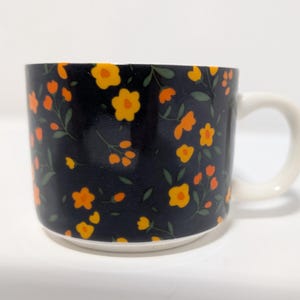 Puede incluir: Una taza de cerámica con asa y base blancas. La taza presenta un fondo azul oscuro con un patrón repetido de flores naranjas y amarillas y hojas verdes. La taza está diseñada para bebidas calientes.