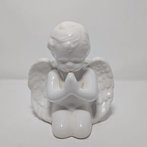 Könnte beinhalten: Eine weiße Keramik-Engelsfigur in kniender Position mit gefalteten Händen. Der Engel hat detaillierte Flügel und trägt ein einfaches Gewand. Die Figur ist ein dekorativer Artikel, der sich zur Präsentation auf einem Regal oder Tisch eignet.