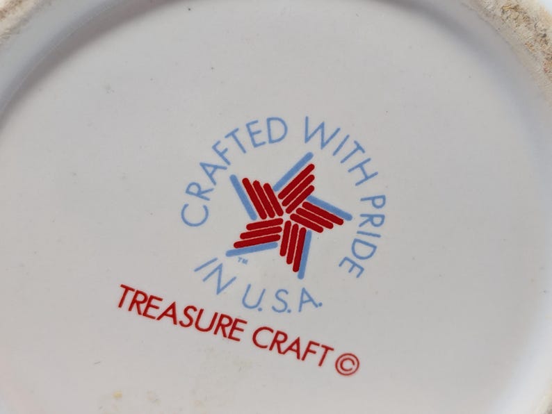 May include: Close-up of a white ceramic surface with a circular emblem. The emblem features a red and blue star design, surrounded by the text "CRAFTED WITH PRIDE IN U.S.A." and "TREASURE CRAFT" in red and blue lettering.