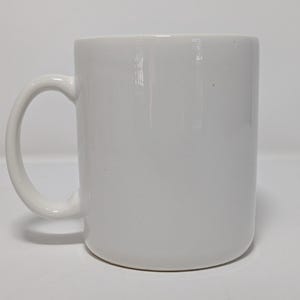 May include: A white ceramic mug with a curved handle. The mug has a cylindrical shape and a smooth, glossy finish. The mug is empty and sits on a white surface.