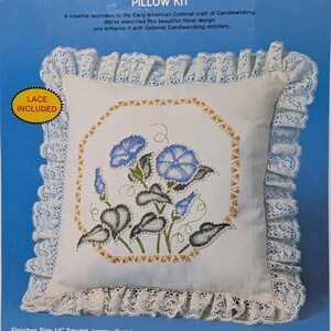 Puede incluir: Un kit de cojín Creative Moments Stencilled Candlewicking que incluye un cojín cuadrado de tela blanca con un diseño floral azul, un borde geométrico amarillo y volantes de encaje blanco. El empaque incluye texto y un código de barras.