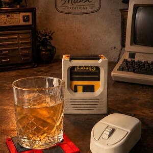 Op de afbeelding: Een helder glas gevuld met een amberkleurige vloeistof op een rood-zwarte diskette-onderzetter, naast een beige computermuis en een vierkante beige houder met meerdere diskettes waarop het woord Cheers! is afgedrukt.