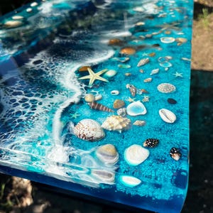 May include: A rectangular table surface featuring a resin art design that mimics an ocean shoreline. The clear blue resin incorporates white wave patterns and a variety of embedded seashells, including starfish, conch, and spiral shells.