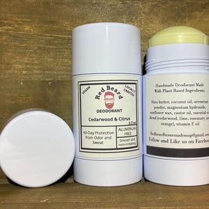May include: Three white deodorant containers. One is open, showing a yellow top. The labels say "Red Beard" and "Cedarwood & Citrus", with details about ingredients and being aluminium-free. The background is a wooden surface.