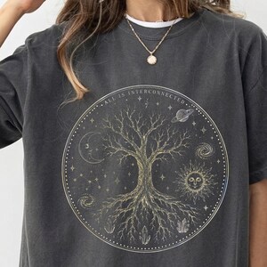 May include: A charcoal gray short-sleeve t-shirt featuring a circular graphic print of a tree with exposed roots. The design includes celestial elements like a sun, moon, stars, and planets, with the text ALL IS INTERCONNECTED encircling the tree.