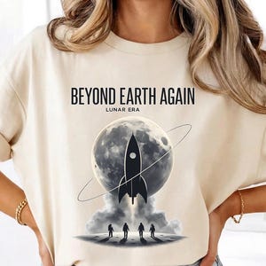 May include: Beige t-shirt with the text "BEYOND EARTH AGAIN" and "LUNAR ERA". The graphic features a black rocket ship in front of a full moon with a ring around it, and four silhouetted figures on a planet surface.