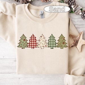 May include: A sand-colored sweatshirt featuring five Christmas tree designs. Each tree has a unique pattern, including floral, plaid, and gingham. The trees are in shades of green, red, and cream. The word "Sand" is printed on the top of the sweatshirt.