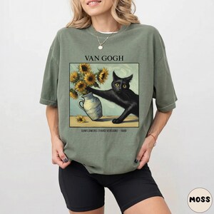 May include: Olive green t-shirt featuring a graphic of a black cat reaching for a vase of sunflowers, inspired by Van Gogh's art. The text "VAN GOGH" and "SUNFLOWERS (THIRD VERSION) - 1888" are printed above and below the image.