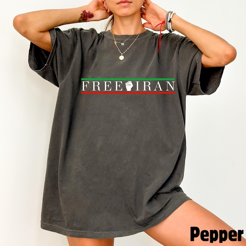 May include: Dark gray t-shirt with the text "FREE IRAN" in white, with a green and red stripe design. The shirt features a raised fist graphic. The word "Pepper" is printed in the bottom right corner.