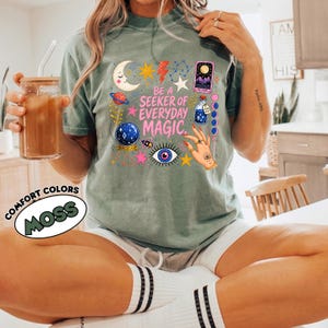 May include: A moss green t-shirt featuring a graphic print with the text BE A SEEKER OF EVERYDAY MAGIC. The design includes a crescent moon, stars, a planet, a tarot card, a potion bottle, a crystal ball, an eye, and a hand with rings.