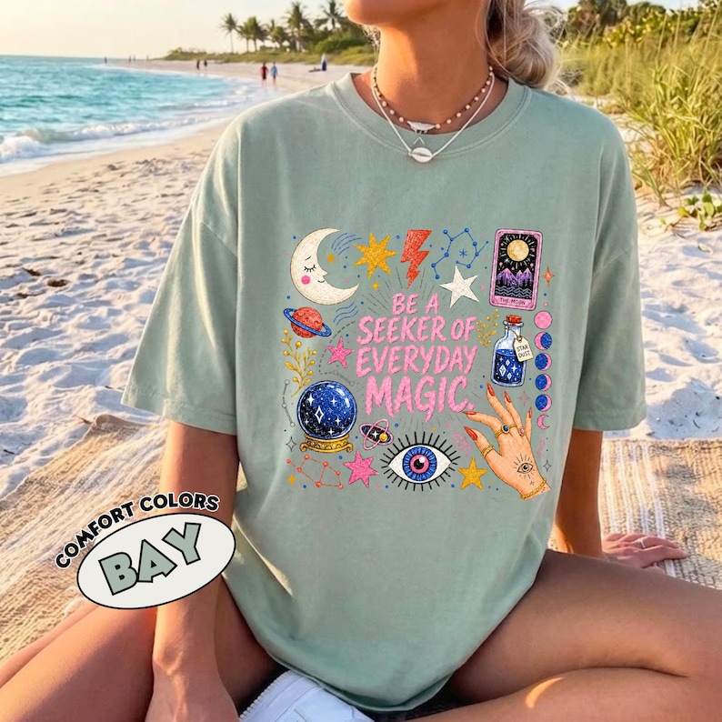 May include: A sage green short-sleeve t-shirt featuring a graphic print with the text BE A SEEKER OF EVERYDAY MAGIC. The design includes a crescent moon, stars, a lightning bolt, a tarot card, a crystal ball, an eye, and a hand with rings.
