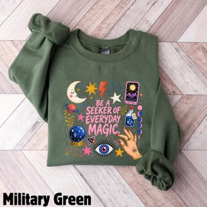 May include: A military green crewneck sweatshirt featuring a graphic print with the text BE A SEEKER OF EVERYDAY MAGIC. The design includes a crescent moon, stars, a planet, a crystal ball, a tarot card, a potion bottle, and a hand with rings.
