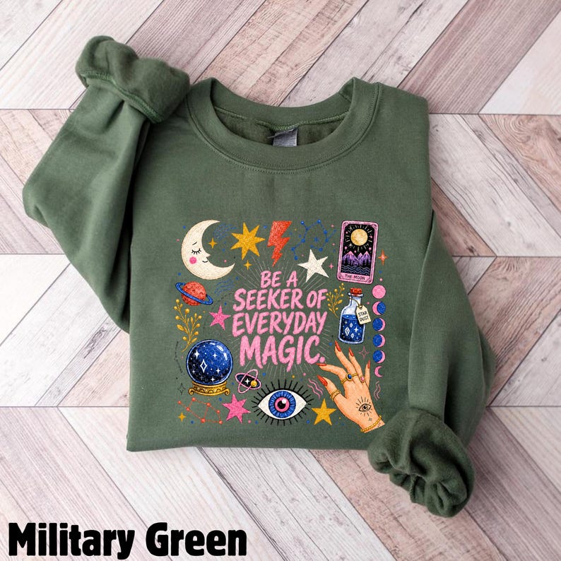 May include: A military green crewneck sweatshirt featuring a graphic print with the text BE A SEEKER OF EVERYDAY MAGIC. The design includes a crescent moon, stars, a planet, a crystal ball, a tarot card, a potion bottle, and a hand with rings.
