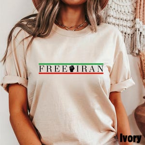 May include: Ivory-colored t-shirt with the text "FREE IRAN" in black, green, and red. The design includes a clenched fist symbol. The shirt has short sleeves and a relaxed fit.