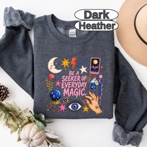 May include: A dark heather grey crewneck sweatshirt featuring a graphic print with the text "BE A SEEKER OF EVERYDAY MAGIC." The design includes a crescent moon, stars, a tarot card, a crystal ball, a potion bottle, and a hand with rings.