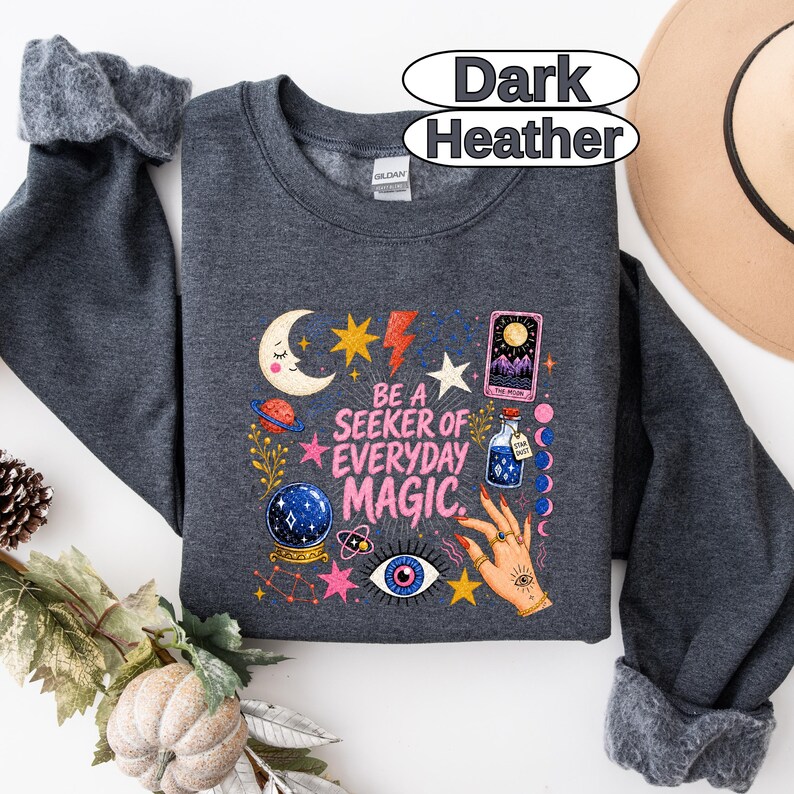 May include: A dark heather grey crewneck sweatshirt featuring a graphic print with the text "BE A SEEKER OF EVERYDAY MAGIC." The design includes a crescent moon, stars, a tarot card, a crystal ball, a potion bottle, and a hand with rings.