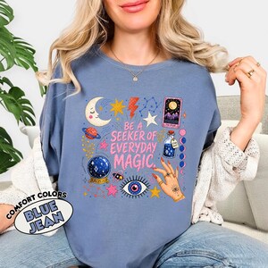 May include: A blue jean coloured crewneck sweatshirt featuring a graphic print with the text BE A SEEKER OF EVERYDAY MAGIC. The design includes a crescent moon, stars, a planet, a crystal ball, a tarot card, a potion bottle, and a hand with rings.