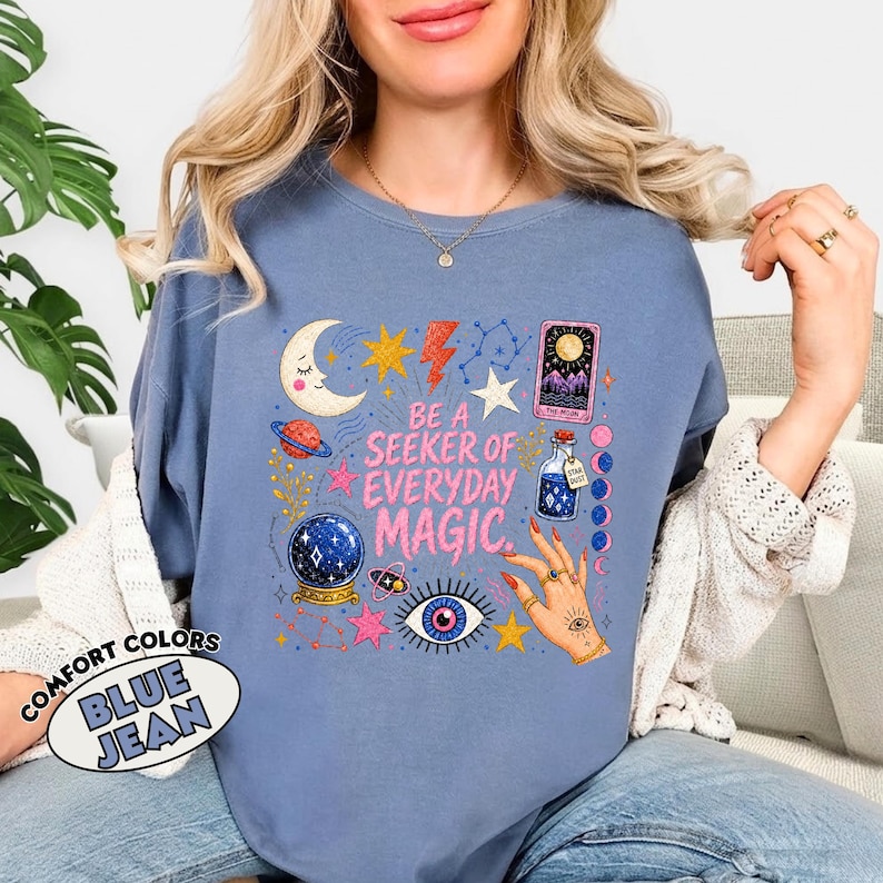 May include: A blue jean coloured crewneck sweatshirt featuring a graphic print with the text BE A SEEKER OF EVERYDAY MAGIC. The design includes a crescent moon, stars, a planet, a crystal ball, a tarot card, a potion bottle, and a hand with rings.