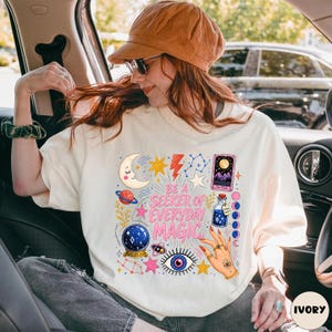 May include: An off-white t-shirt featuring a colourful graphic print with the text BE A SEEKER OF EVERYDAY MAGIC. The design includes a crescent moon, stars, a planet, a crystal ball, a tarot card, a potion bottle, and a hand with an eye symbol.