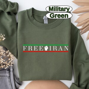 May include: Military green sweatshirt with the text "FREE IRAN" in white, with a red and green stripe. The design includes a white fist. The sweatshirt is folded.