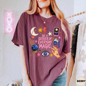 May include: A berry-coloured short-sleeve t-shirt featuring a graphic print with the text BE A SEEKER OF EVERYDAY MAGIC. The design includes a crescent moon, stars, a lightning bolt, a tarot card, a crystal ball, a potion bottle, and an eye.