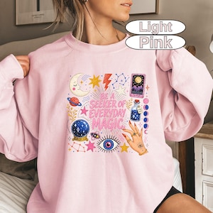 May include: A light pink sweatshirt featuring a colourful graphic print with the text "BE A SEEKER OF EVERYDAY MAGIC." The design includes a crescent moon, stars, a lightning bolt, a tarot card, a potion bottle, a crystal ball, and an eye.