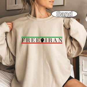 May include: Sand-colored sweatshirt with the text "FREE IRAN" in black, green, and red. The design includes a black fist graphic. The sweatshirt is a long-sleeved crew neck.