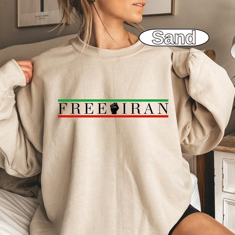 May include: Sand-colored sweatshirt with the text "FREE IRAN" in black, green, and red. The design includes a black fist graphic. The sweatshirt is a long-sleeved crew neck.
