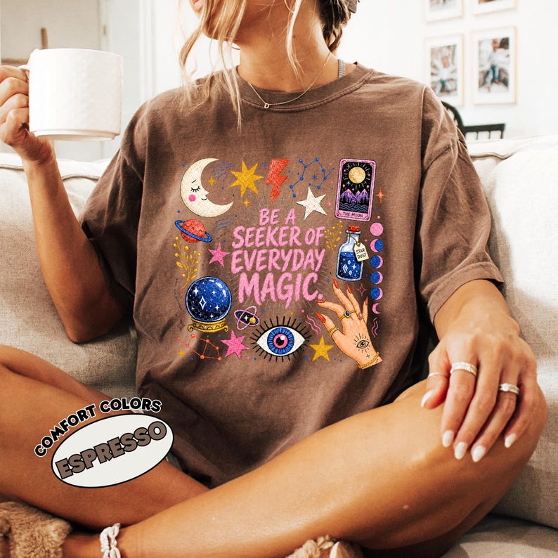 May include: A brown short-sleeve t-shirt featuring a colourful graphic print with the text BE A SEEKER OF EVERYDAY MAGIC. The design includes a crescent moon, stars, a planet, a crystal ball, a tarot card, a potion bottle, and a hand with rings.