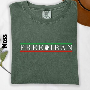 May include: Moss green t-shirt with the text "FREE IRAN" in white, with a green and red stripe design. The shirt has a crew neck and a Comfort Colors tag.
