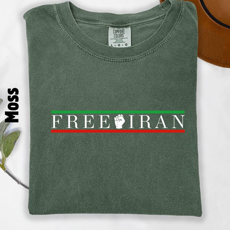 May include: Moss green t-shirt with the text "FREE IRAN" in white, with a green and red stripe design. The shirt has a crew neck and a Comfort Colors tag.