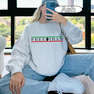 May include: Ash-colored sweatshirt with the text "FREE IRAN" in green and black, with a red and green stripe. The sweatshirt is paired with light blue jeans and white sneakers.