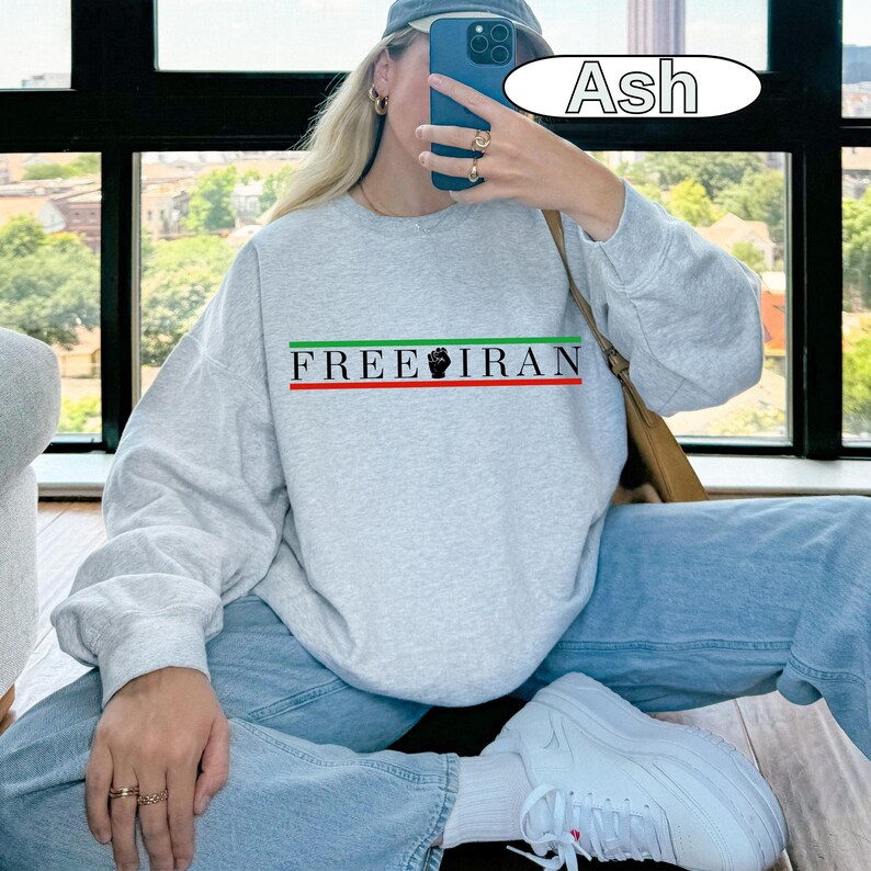 May include: Ash-colored sweatshirt with the text "FREE IRAN" in green and black, with a red and green stripe. The sweatshirt is paired with light blue jeans and white sneakers.