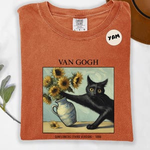 May include: Orange Comfort Colors t-shirt featuring a black cat reaching for a vase of sunflowers, a parody of Van Gogh's painting. The text "VAN GOGH" and "SUNFLOWERS (THIRD VERSION) 1888" are printed on the shirt. A "YAM" sticker is on the shirt.