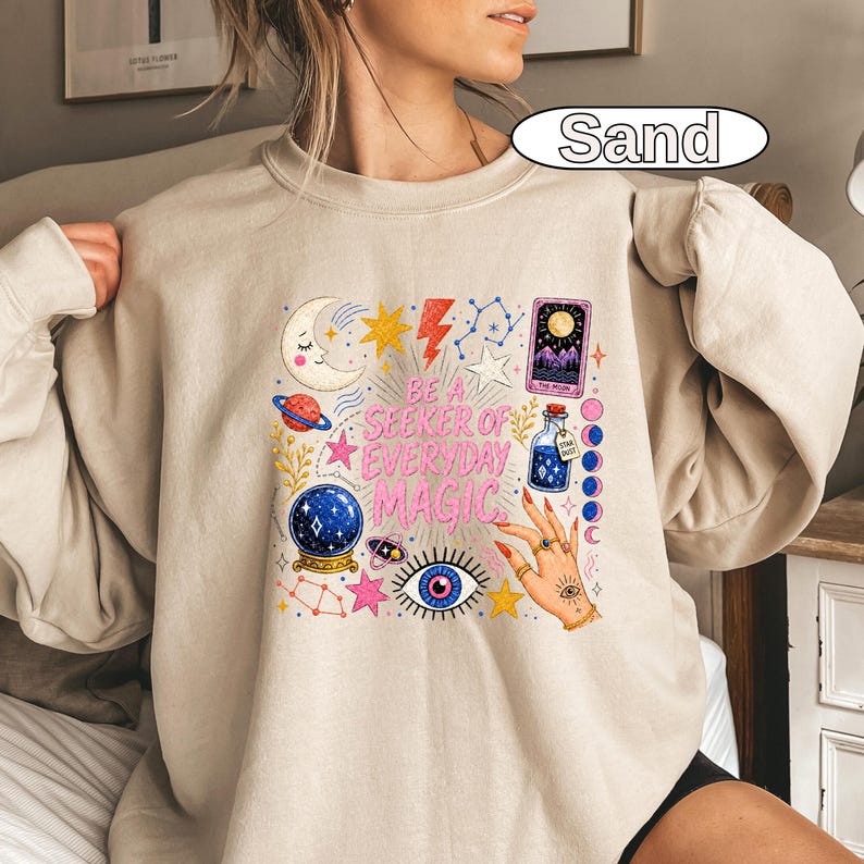 May include: A sand-coloured crewneck sweatshirt featuring a graphic print with the text "BE A SEEKER OF EVERYDAY MAGIC." The design includes a crescent moon, stars, a lightning bolt, a tarot card, a potion bottle, a crystal ball, and an eye.