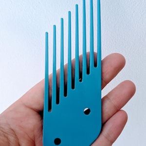 May include: A teal hair pick with long, evenly spaced teeth. The pick has a rounded base with two circular holes. The teeth are of varying lengths, creating a stepped appearance. The comb is held in a hand against a white background.