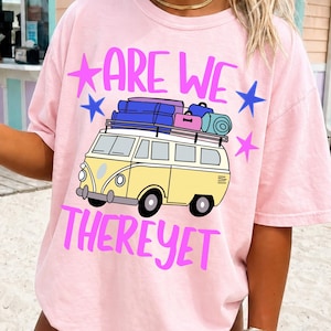 Op de afbeelding: Lichtroze T-shirt met een gele busje-afbeelding beladen met bagage. De tekst "ARE WE" en "THERE YET" zijn paars, met blauwe sterren. Het shirt heeft een relaxte, oversized pasvorm.