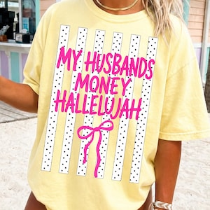 May include: A pale yellow short-sleeve t-shirt featuring a graphic print of vertical white stripes with black dots. The text reads MY HUSBANDS MONEY HALLELUJAH in bright pink letters, accompanied by a pink line-drawn bow graphic centered below.