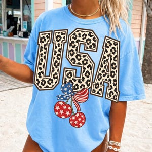 Può includere: T-shirt azzurra con la scritta "USA" in un motivo leopardato. Sotto il testo c'è una grafica di ciliegie con un fiocco rosso, bianco e blu. La maglietta è in stile casual.
