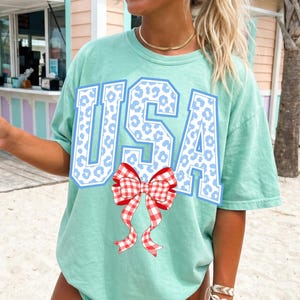 Op de afbeelding: Lichtgroen T-shirt met het woord "USA" in blauwe letters en een wit luipaardprintpatroon. Een rood-witte ruitstrik is eronder gecentreerd. De persoon draagt het shirt op het strand.
