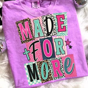 May include: A lilac t-shirt with the slogan "MADE FOR MORE" in a colourful patchwork design. The text incorporates leopard print, stripes, and polka dots. The shirt is a Comfort Colors brand.