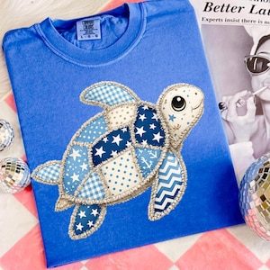 May include: A blue Comfort Colors t-shirt featuring a patchwork turtle design. The turtle has blue and white patterned patches with stars, dots, and anchors. The shirt is displayed with a newspaper and disco balls.