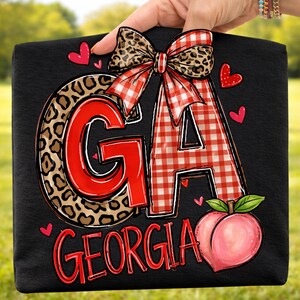 Puede incluir: Bolso tote negro con las letras "GA" en rojo y estampado de leopardo, un lazo de cuadros y un gr&aacute;fico de melocot&oacute;n. La palabra "GEORGIA" est&aacute; escrita en rojo debajo. Peque&ntilde;os corazones rojos est&aacute;n dispersos alrededor del dise&ntilde;o.