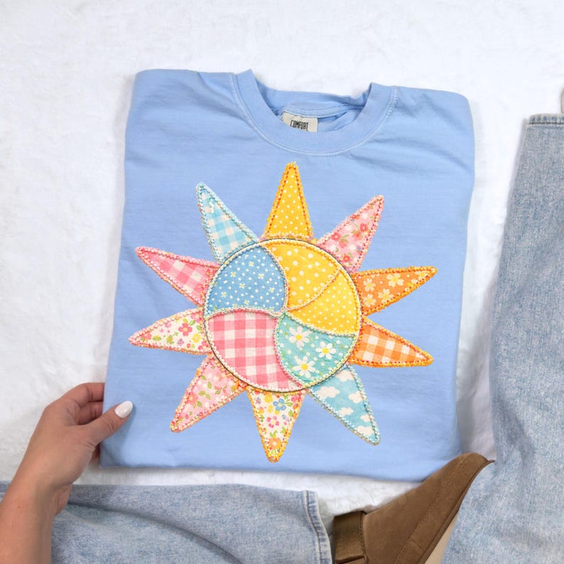 Puede incluir: Una sudadera azul claro con un aplique de sol colorido. El dise&ntilde;o del sol presenta varias telas estampadas, incluyendo cuadros, flores y lunares, en tonos de rosa, amarillo, naranja y azul. La sudadera est&aacute; doblada y colocada sobre una superficie blanca.