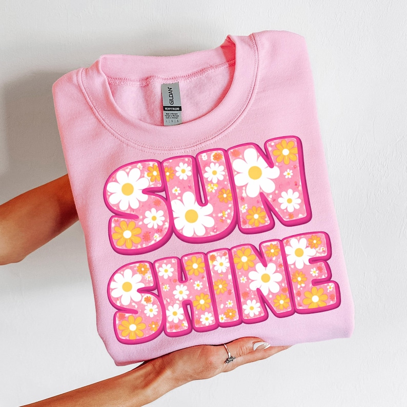 Pu&ograve; includere: Felpa rosa chiaro con la scritta "SUNSHINE" in grandi lettere a bolla. Le lettere sono riempite con un motivo floreale di margherite bianche e gialle. La felpa &egrave; piegata.