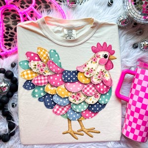 Puede incluir: Camiseta color crema con un diseño de gallina de patchwork colorido. La gallina tiene una cresta rosa, un pico amarillo y un cuerpo hecho de varias telas estampadas, incluyendo lunares, cuadros y estampados florales. Un vaso a cuadros rosa y blanco está a la derecha.