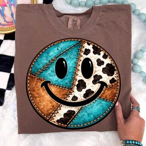 May include: A brown Comfort Colors t-shirt featuring a large smiley face design. The smiley face is divided into sections of turquoise, brown, and cow print, stitched together with visible seams. The design has black eyes and a curved black smile.