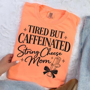 May include: A coral Comfort Colors t-shirt with the words "TIRED BUT CAFFEINATED String Cheese Mom" in black script. A graphic of string cheese tied with a bow is below the text. The shirt is size XL-TG-80.
